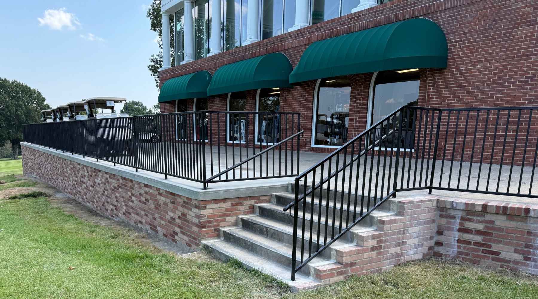 A brick building with green awnings has a raised concrete patio with a black metal railing and steps leading down to a grassy area. Golf carts are parked to the left of the building.