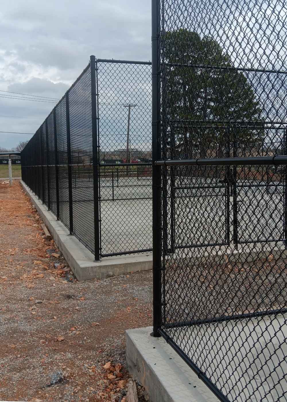 A tall black chain-link fence surrounds a concrete area under a cloudy sky, with trees and utility poles visible in the background. The ground outside the fence is covered in gravel and dirt.