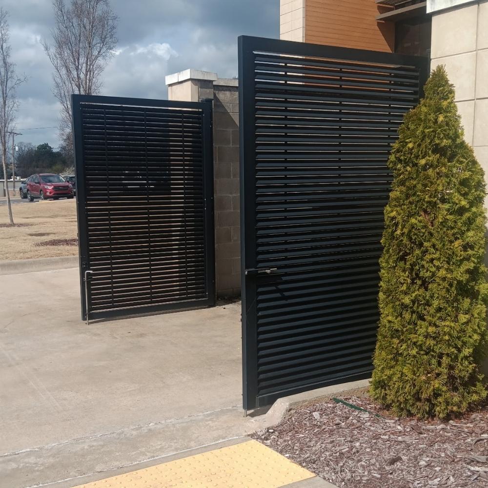 A large black metal gate with horizontal slats stands open next to a smaller matching gate. The gates are next to a tan building and a bush, with parked cars and trees visible in the background.
