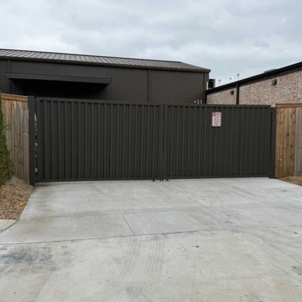 A large, closed metal gate blocks entry to a paved driveway, flanked by wooden fences on both sides. Two buildings are visible behind the gate under a cloudy sky.