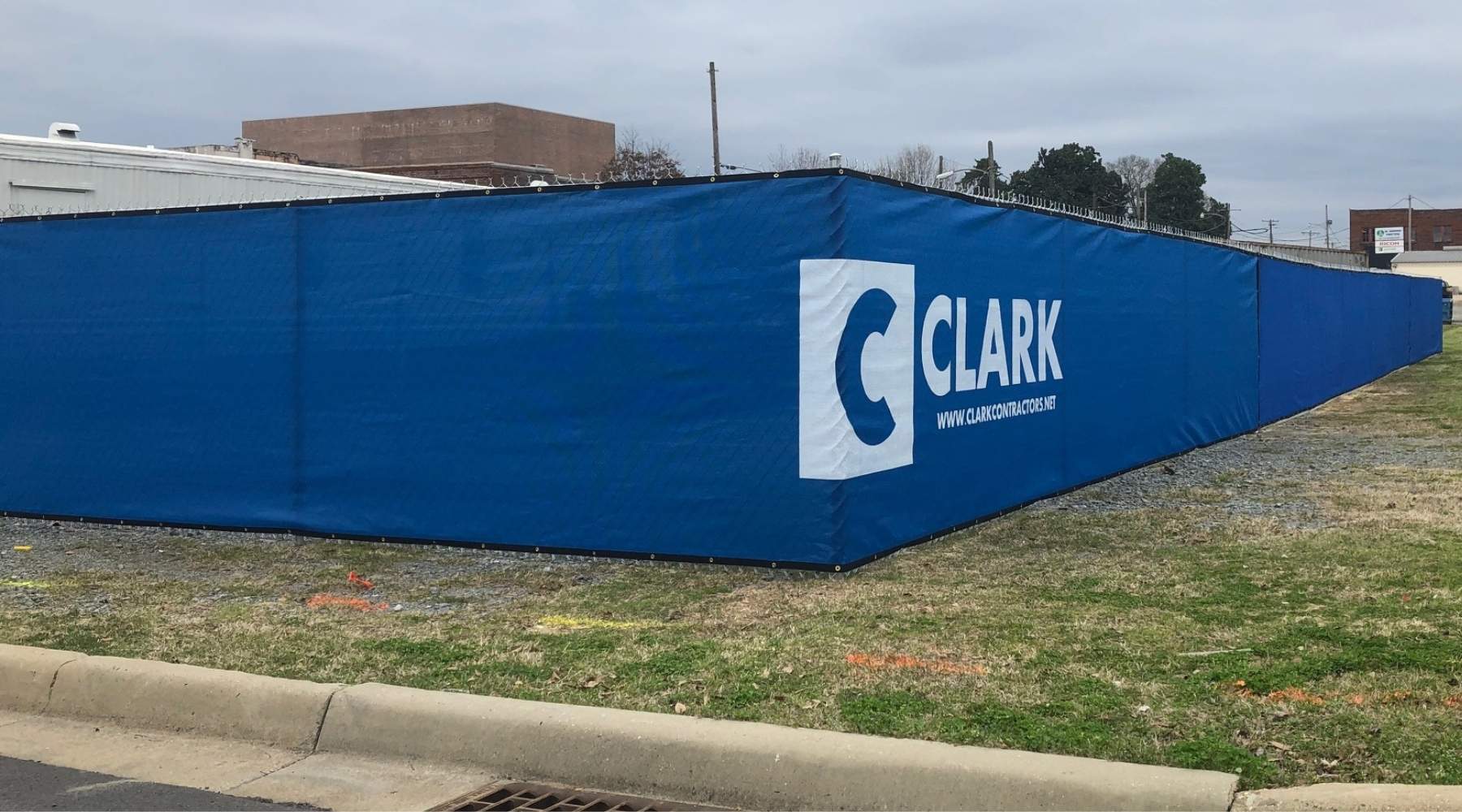 A blue construction fence with the white Clark Construction logo and website printed on it surrounds a grassy area near a street curb. Industrial buildings and trees are visible in the background.