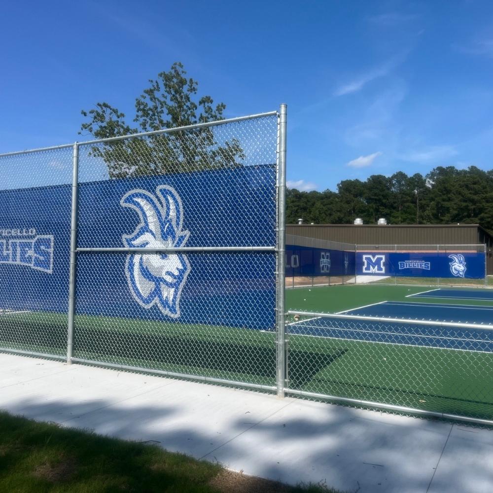 A fenced outdoor tennis court features a large blue windscreen with a white outline of a goats head and Monticello Billies logo, alongside trees and a blue sky in the background.
