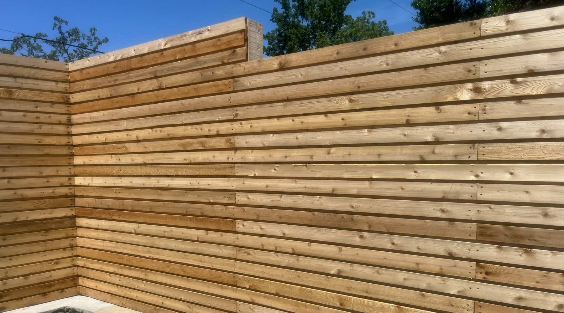 A wooden privacy fence made of horizontal slats forms a corner under a clear blue sky, with green trees visible above the fence line.