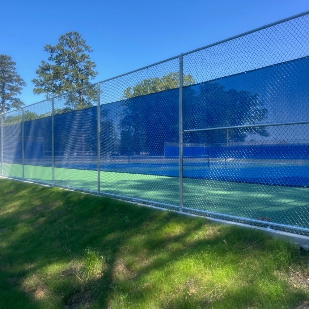A tennis court with blue windscreens on a chain-link fence, surrounded by green grass and trees, under a clear blue sky.