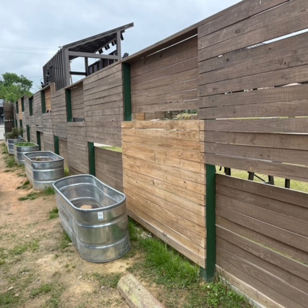 Several large metal tubs are lined up beside a tall wooden fence made of horizontal planks, with some sections lighter than others. The ground is dirt and grass, and the sky is overcast.