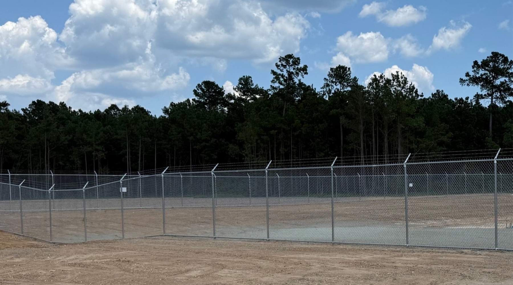 A fenced, barren dirt lot with barbed wire on top, surrounded by tall trees and a forested area under a blue sky with scattered clouds.