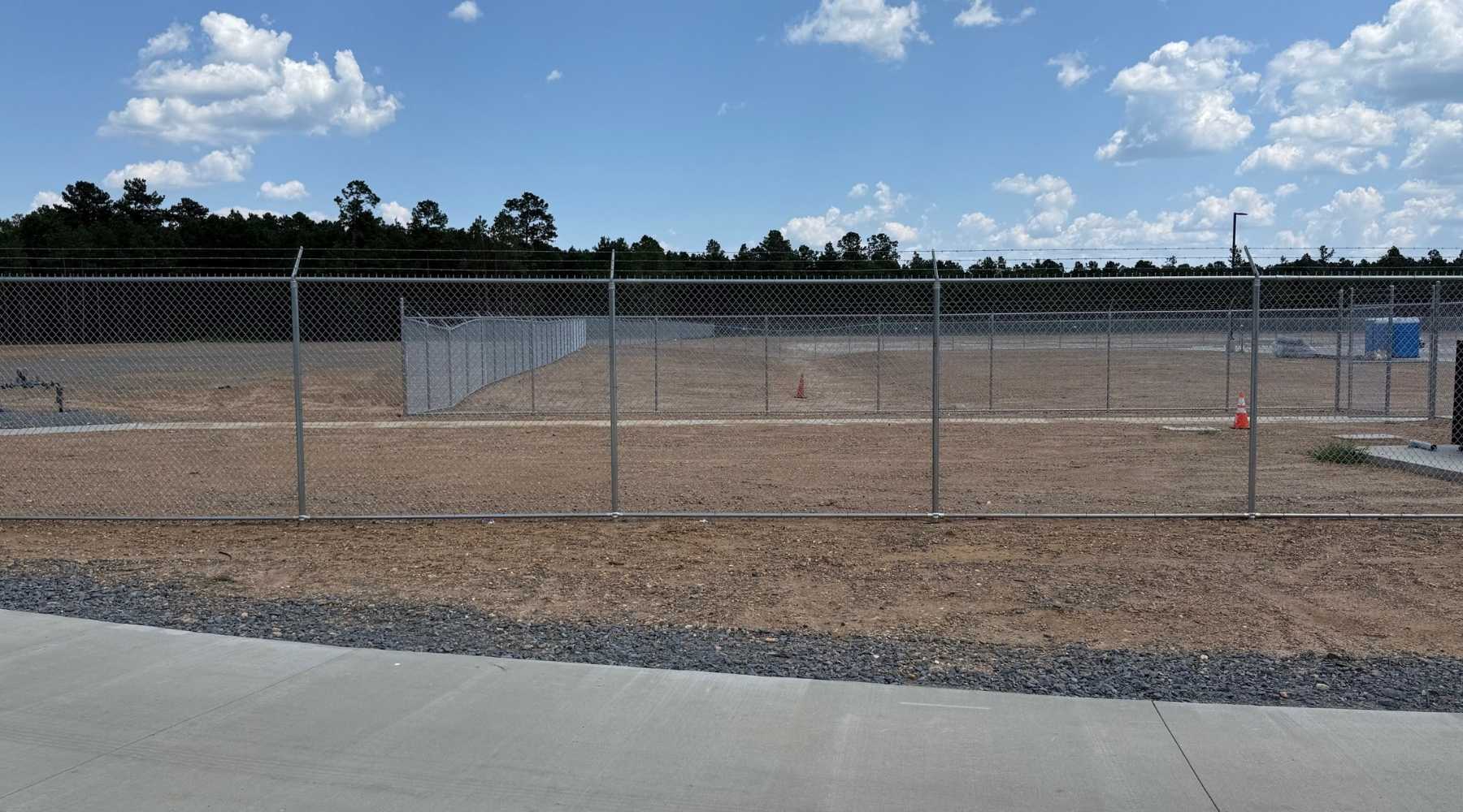 A fenced, empty dirt lot with a few orange traffic cones sits under a blue sky with scattered clouds. Trees line the background beyond the fence, and part of a paved sidewalk is visible in the foreground.