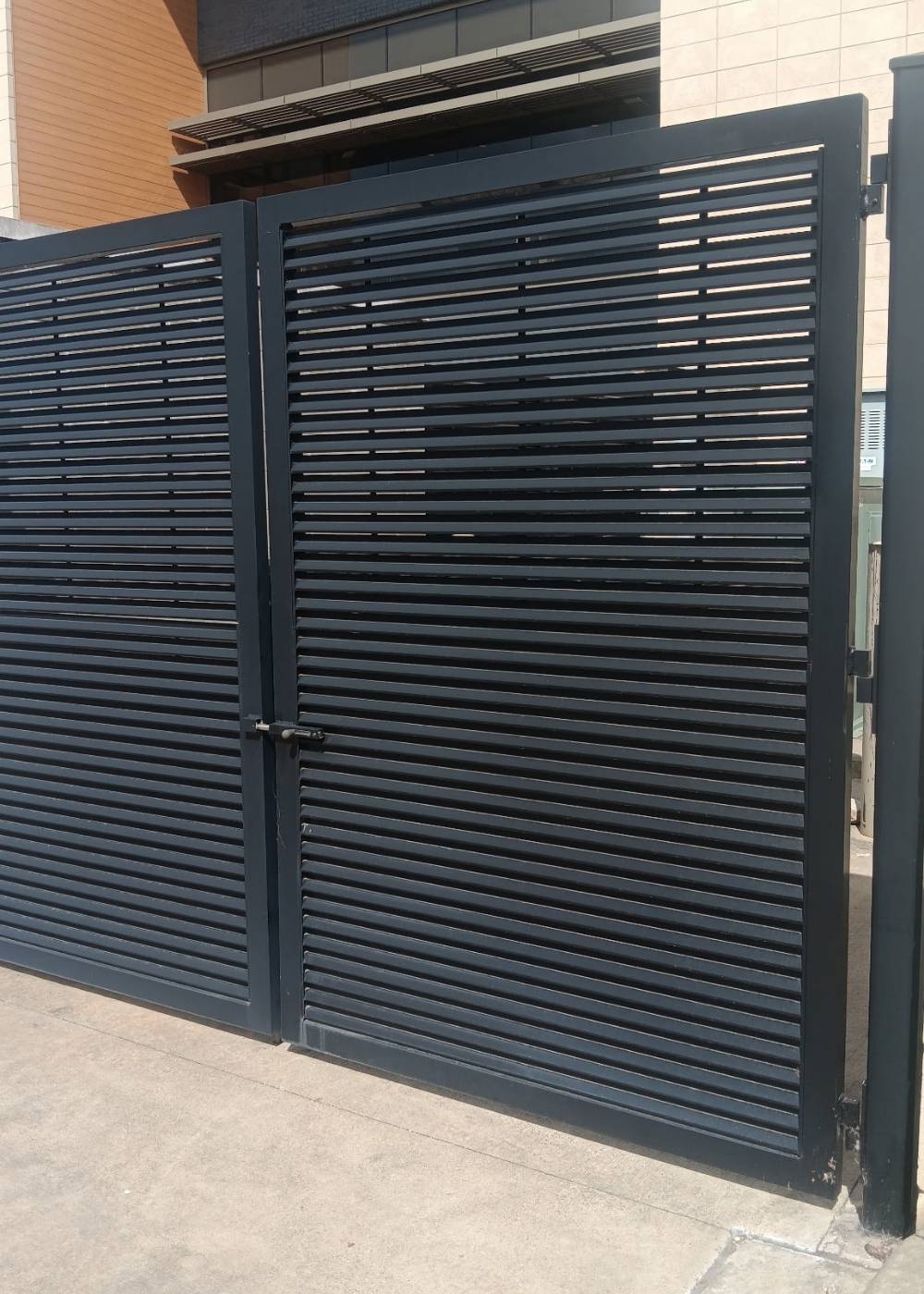 A large black metal gate with horizontal slats is partially open, standing on a concrete surface in front of a modern building with beige and brown walls.