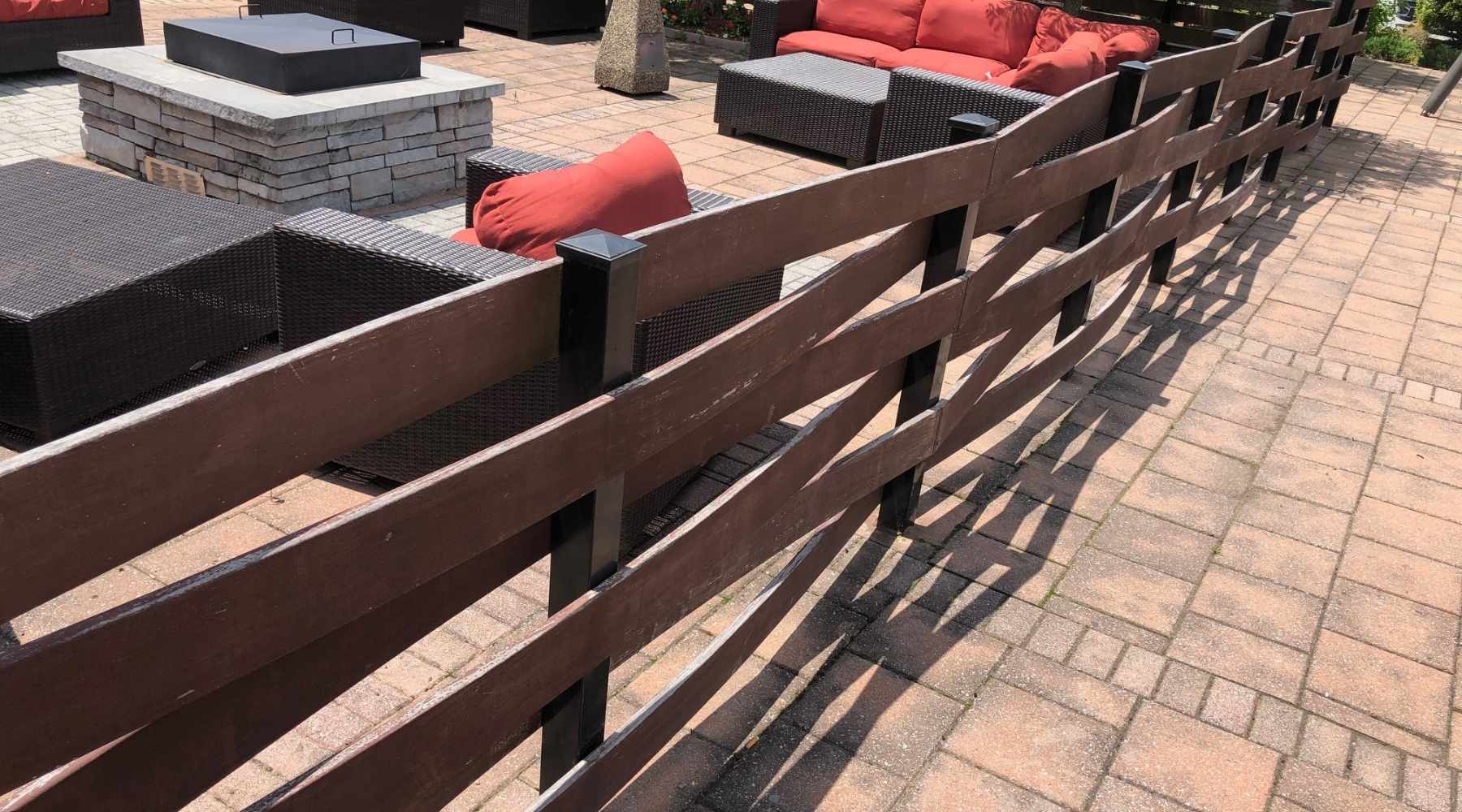 A brown wooden fence runs diagonally across a patio with tan pavers, next to outdoor seating with red cushions and a large fire pit. Shadows from the fence fall across the ground.