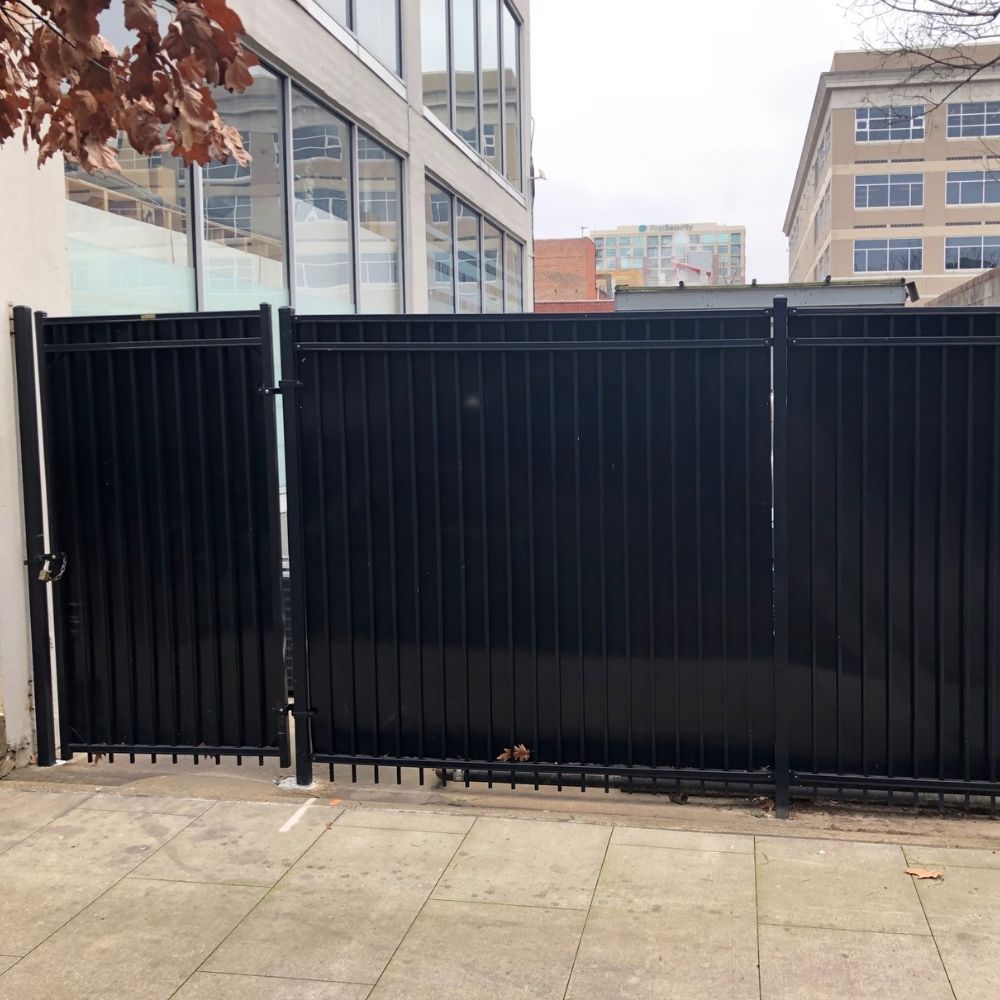 A tall, solid black metal fence blocks a sidewalk in front of modern office buildings; fallen leaves are scattered at the base of the fence.