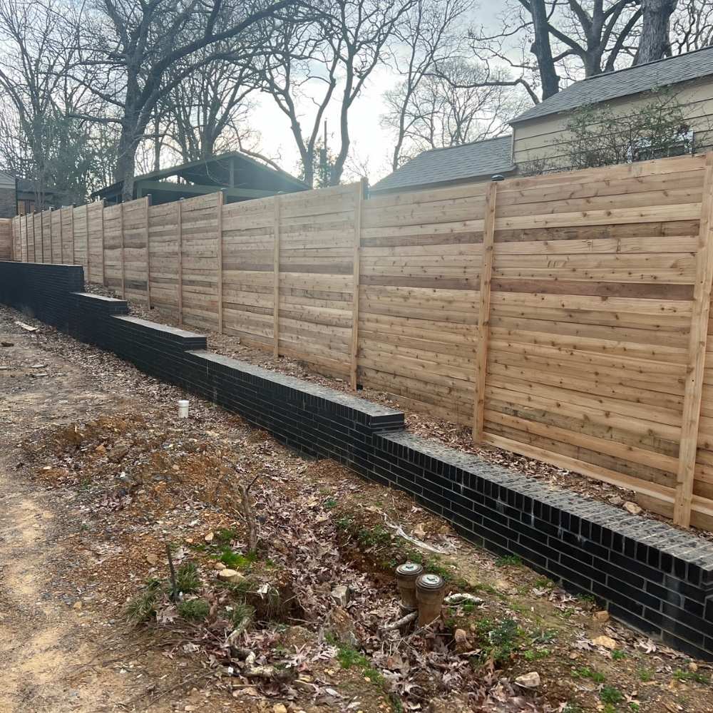 A wooden privacy fence with horizontal planks stands atop a short retaining wall made of dark bricks, bordering a dirt and leaf-covered yard beside a residential area.