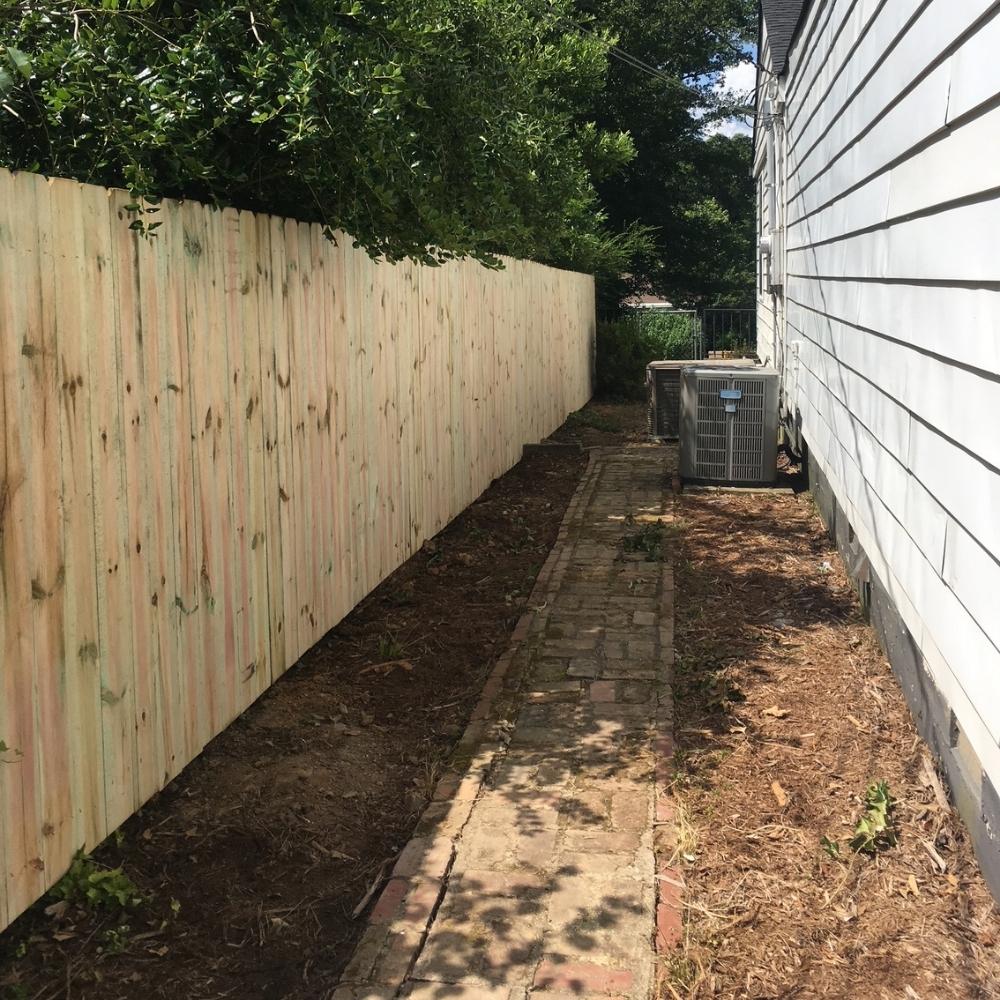 A narrow brick pathway runs between a white wooden house and a tall, light-colored wooden fence, with air conditioning units near the house and trees looming overhead.