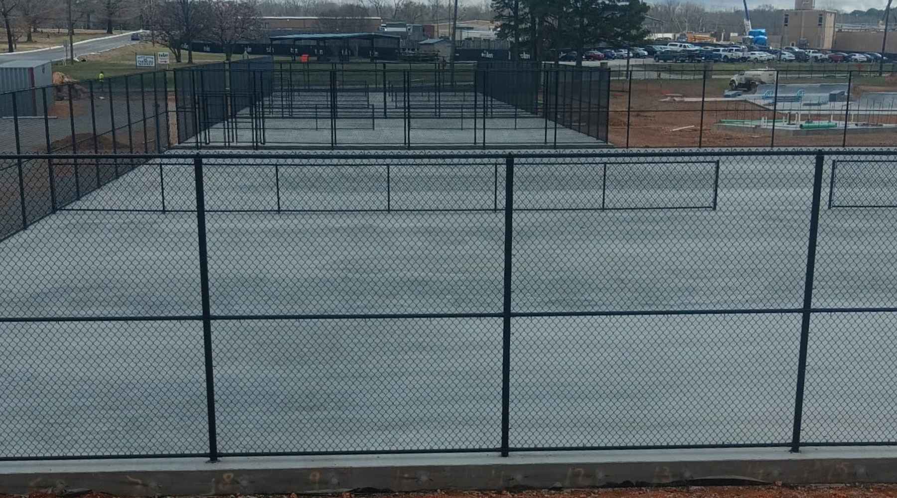 A view of several fenced, empty concrete courts, likely for tennis or pickleball, under overcast skies, with construction materials and a parking lot in the background.