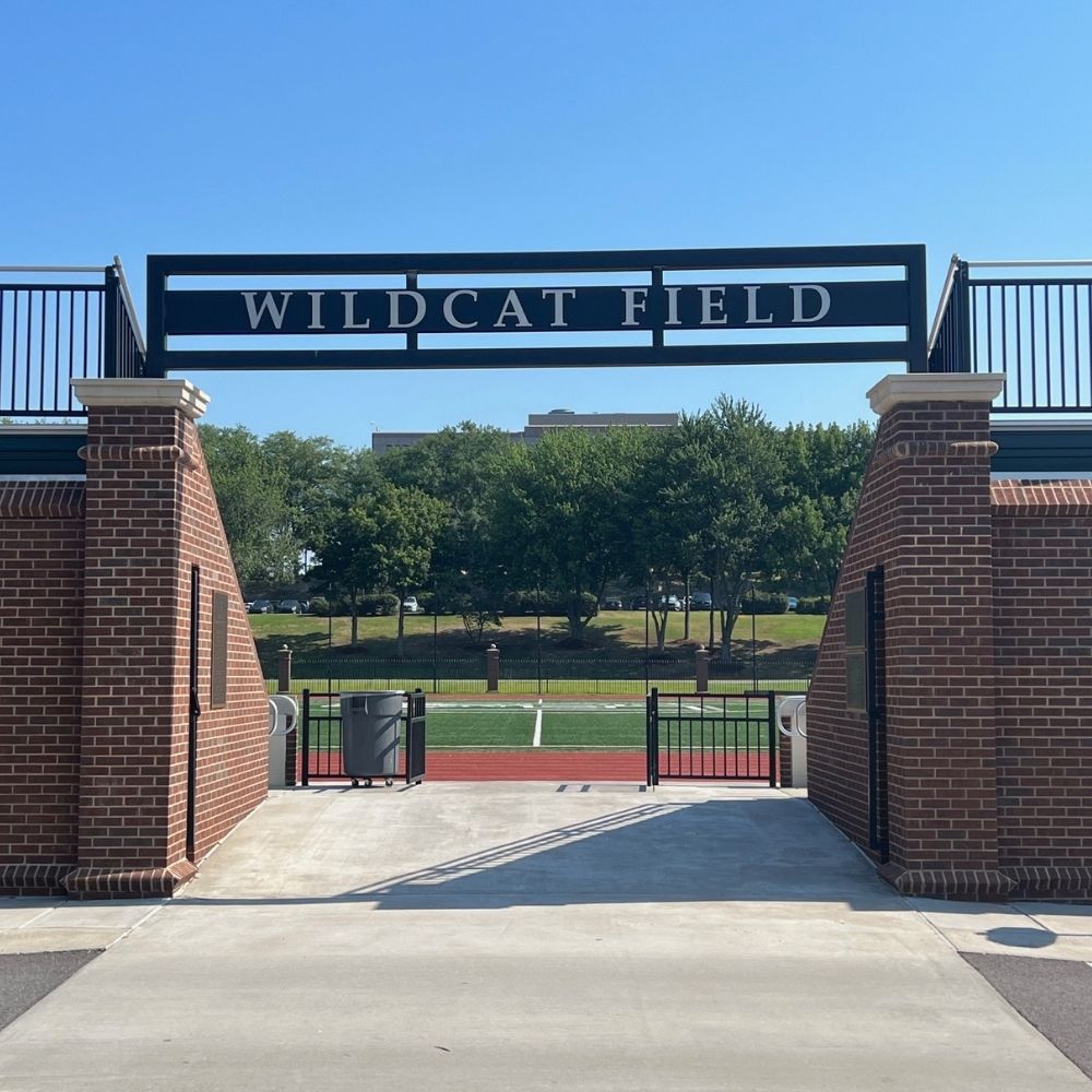 Entrance to a sports field with a sign reading Wildcat Field above brick pillars, leading to a green field and trees in the background under a clear blue sky.