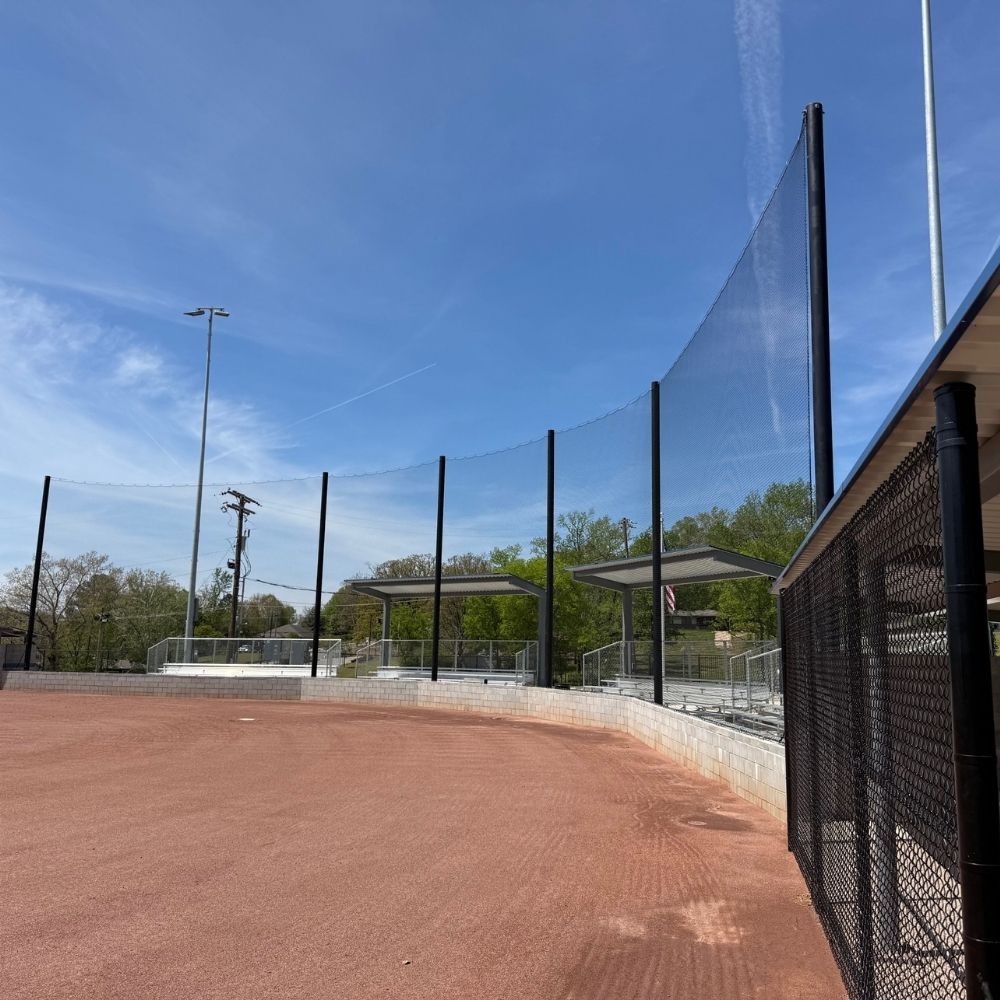 A view from a baseball field looking toward empty bleachers behind a tall net fence under a clear blue sky with some wispy clouds. Trees with green leaves are visible in the background.