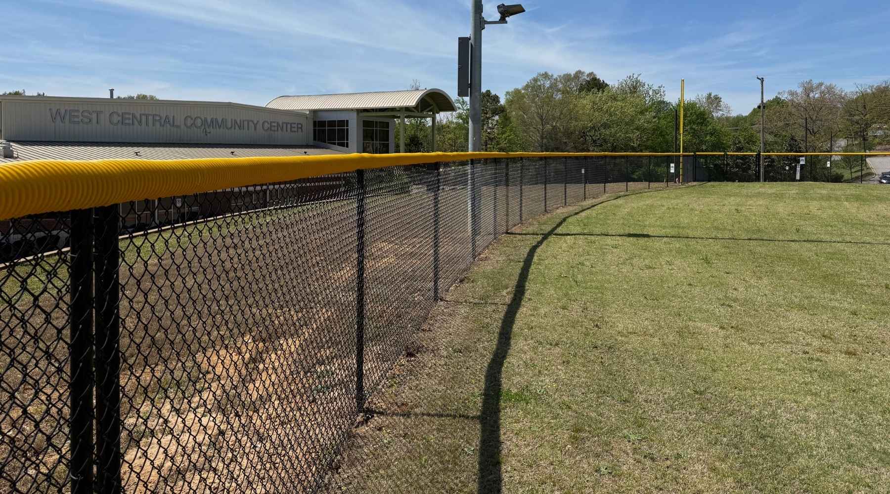A chain-link fence with a yellow guard surrounds a grassy baseball field near the West Central Community Center building under a blue sky.