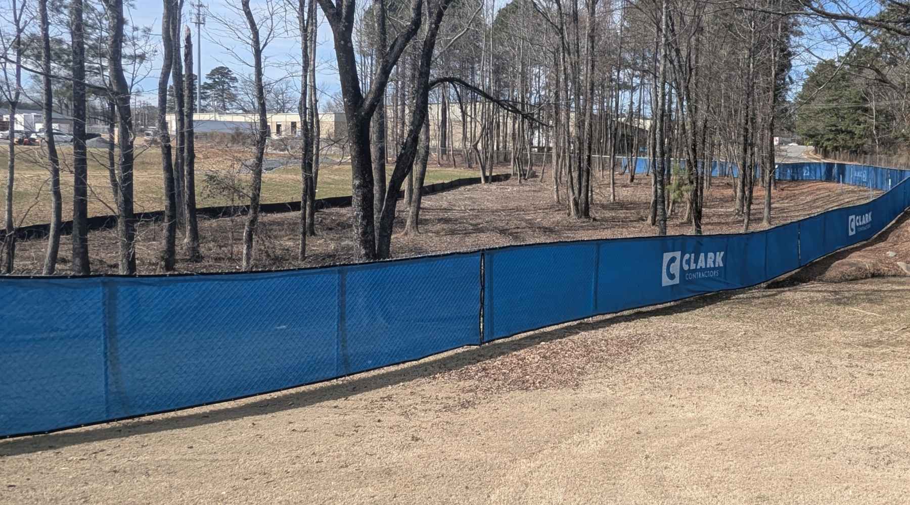 A blue fence labeled Clark Construction runs along the edge of a dirt path through a wooded area with leafless trees on a sunny day.