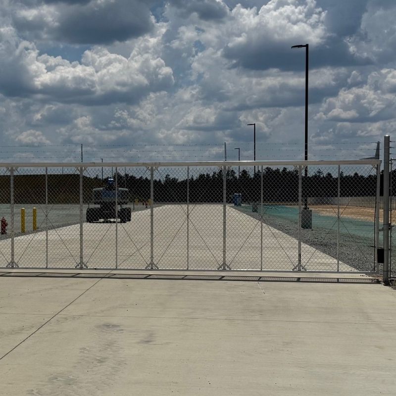 A chain-link gate blocks a wide paved road under a cloudy sky. Street lamps line the road, and trees are visible in the distance. A vehicle is parked behind the gate.