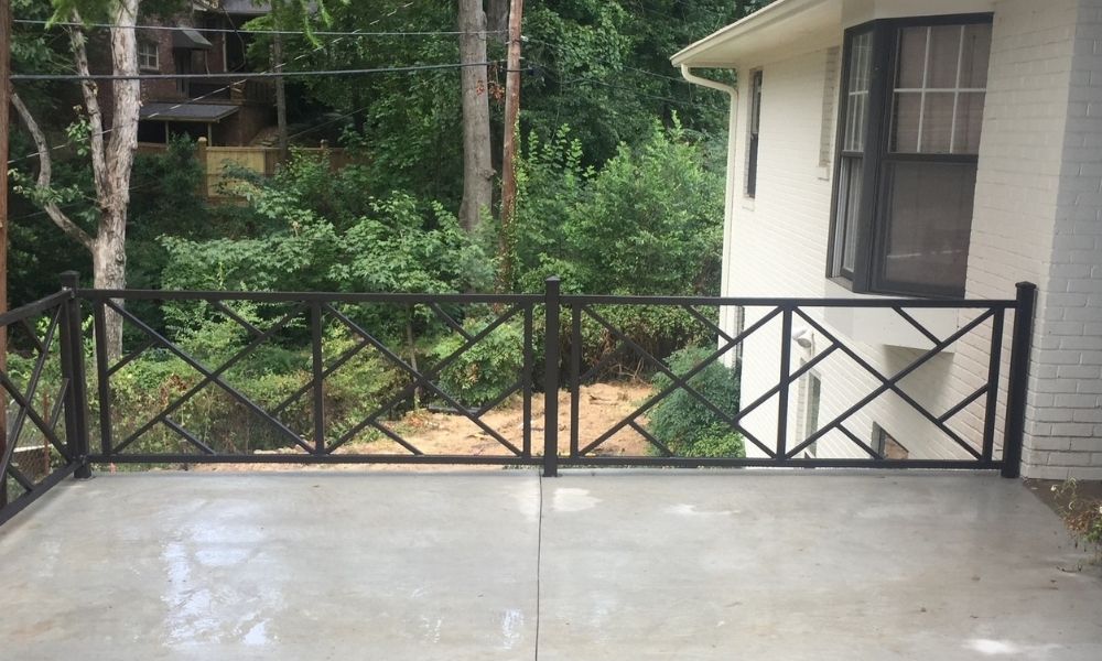 A wet concrete patio with a black metal railing overlooks a wooded area with dense green trees and bushes. A white building is visible on the right, and houses can be seen through the trees in the background.