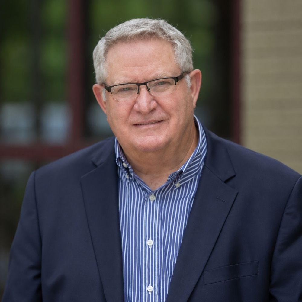 Fence World Harold An older man with gray hair and glasses is wearing a navy blazer over a blue striped shirt, standing outdoors in front of a blurred background of greenery and a brick wall.