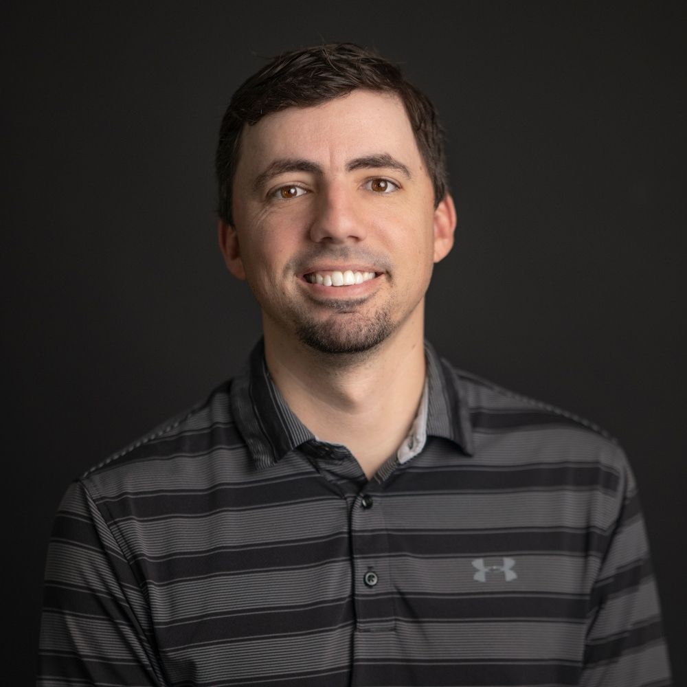 Fence World Luke A man with short dark hair and a trimmed beard smiles at the camera. He is wearing a dark gray, striped polo shirt with the Under Armour logo on the left side. The background is plain black.
